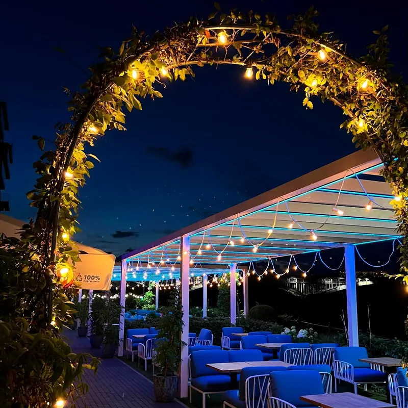 Taras restauracyjny nocą: łuk z roślin z żarówkami girlandy, pergola z niebieskim podświetleniem LED listew i girlandami nad stolikami